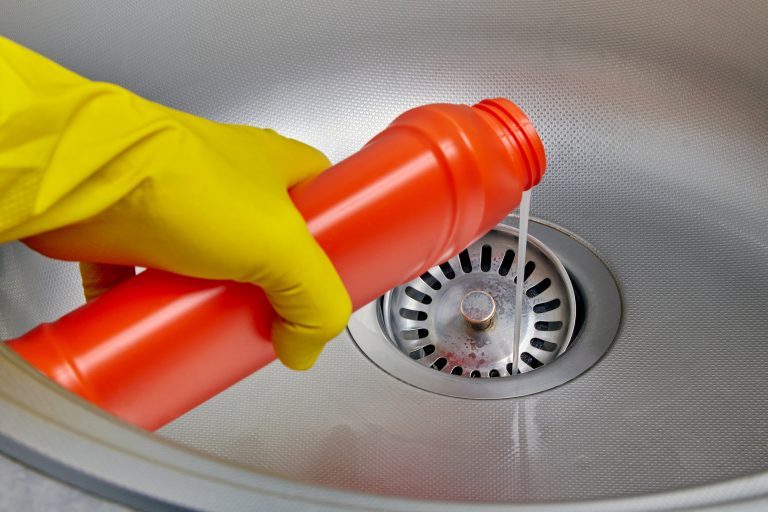 A rubber-gloved hand pouring a chemical drain cleaner into a kitchen sink drain.