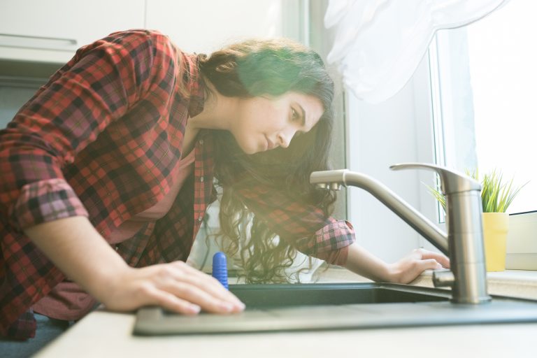 A woman leaning over her kitchen sink to investigate a rotten egg smell coming from the drain.